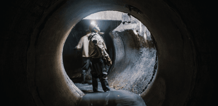 Two drain engineers stand ankle-deep in water in the interior of a sewer drain in Ashford. One of the engineers looks upward towards natural light.