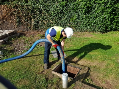 A man in a hard hat and high vis vest lowers a large, flexible blue pipe down a drain surrounded by grass.