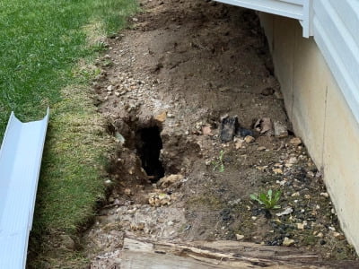 Landscape damage and soil erosion caused by leaking gutters. The grass has receded from the property, leaving unkempt soil and a large hole in the ground.